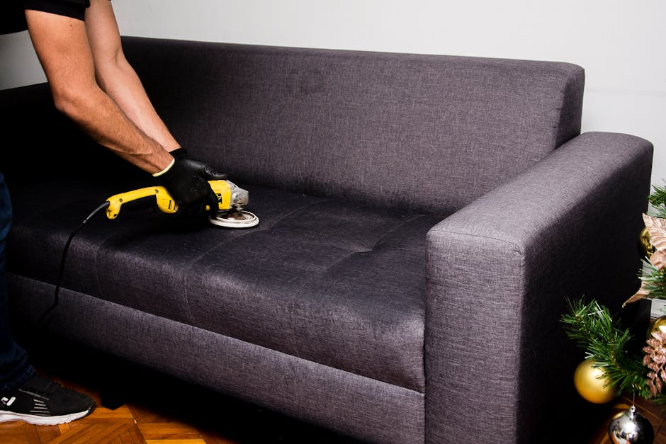 A person wearing black gloves uses a handheld electric upholstery cleaner to deep clean a dark grey fabric sofa in a residential living room. The sofa is positioned against a white wall, with a small 