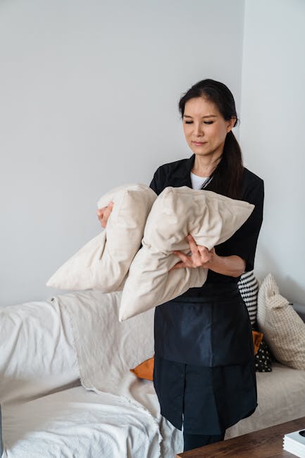 A person wearing black gloves uses a handheld electric upholstery cleaner to deep clean a dark grey fabric sofa in a residential living room. The sofa is positioned against a white wall, with a small 