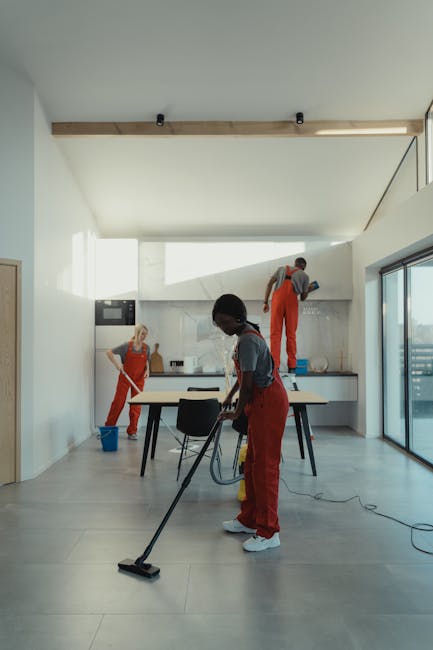 A woman dressed in a grey T-shirt and red overalls is engaged in deep cleaning a modern kitchen. She is holding a mop and standing next to a blue cleaning bucket placed on the light grey tiled floor. 